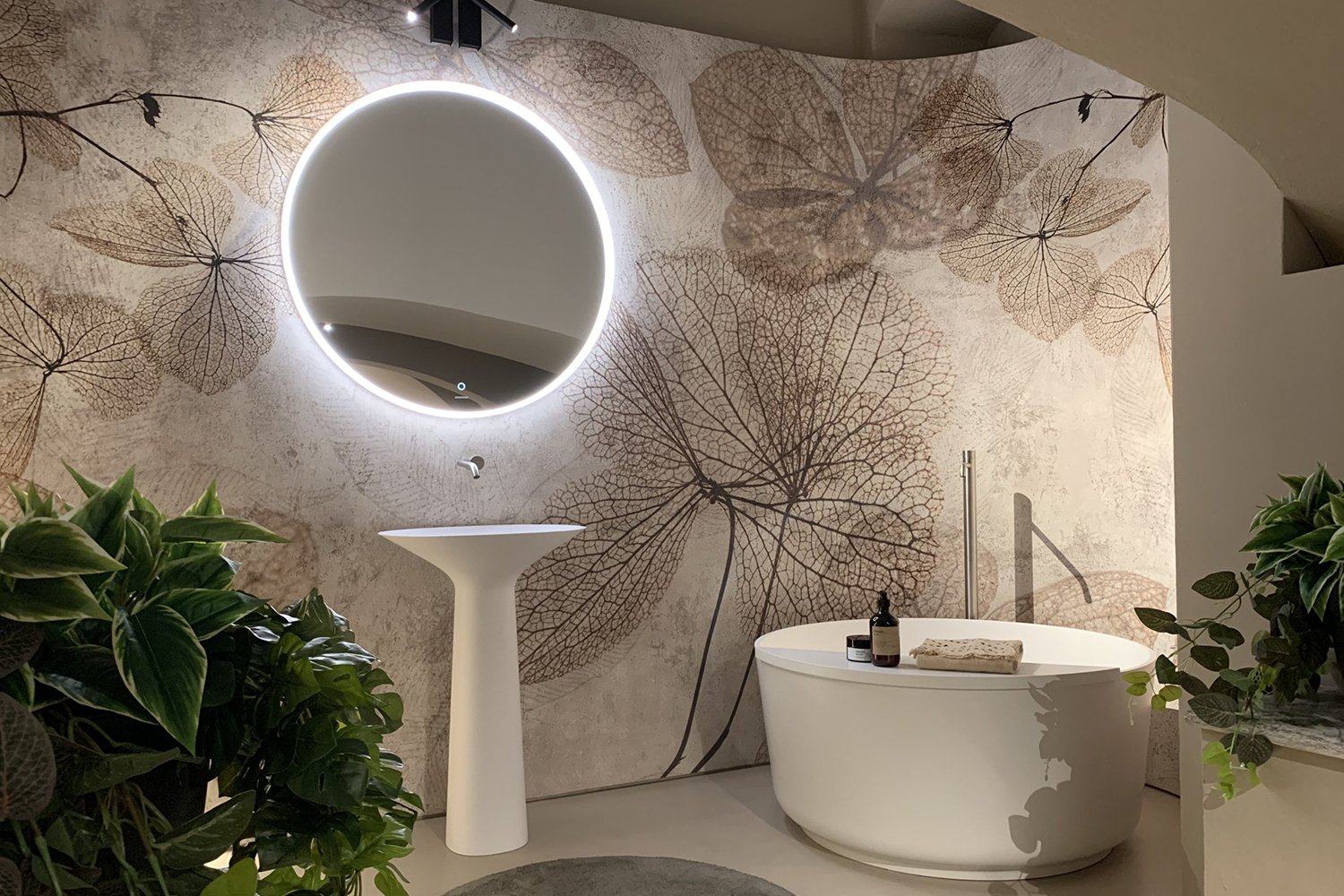 Un angolo interno con carta da parati sui toni del beige e decorazioni floreali. Su di essa emerge uno specchio tondo con cornice a led, un lavandino dal design moderno e una vasca da bagno tonda, moderna ed elegante.